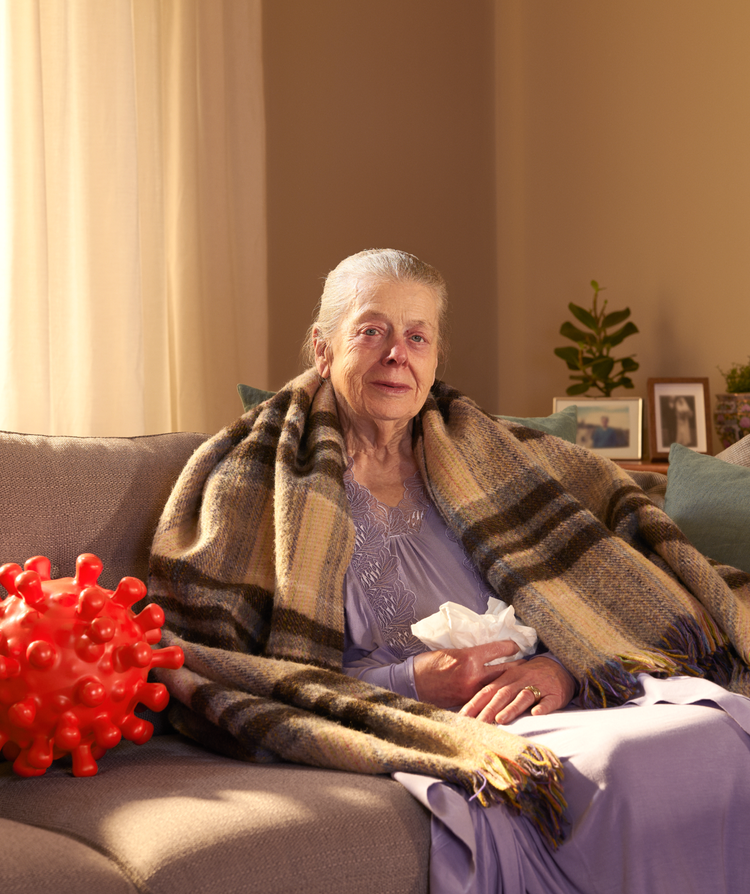 Elderly woman sitting on a sofa wrapped in a blanket with a red nose and tissues in her hand, next to a large red virus molecule