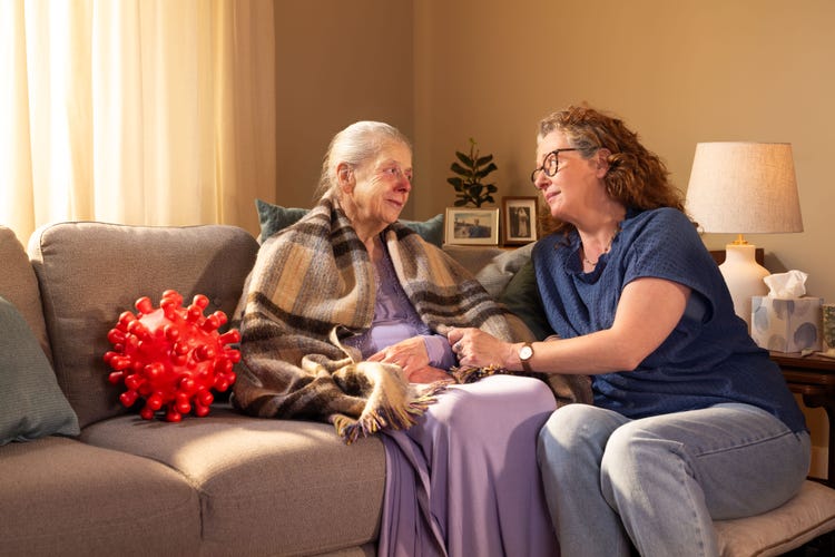 Elderly woman sitting on a sofa wrapped in a blanket with a red nose and next to a large red virus molecule. A younger woman is sitting next to the elderly lady, with her hand on her arm, looking into her eyes.