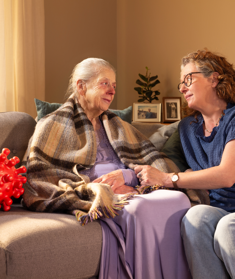 Elderly woman sitting on a sofa wrapped in a blanket with a red nose and next to a large red virus molecule. A younger woman is sitting next to the elderly lady, with her hand on her arm, looking into her eyes.